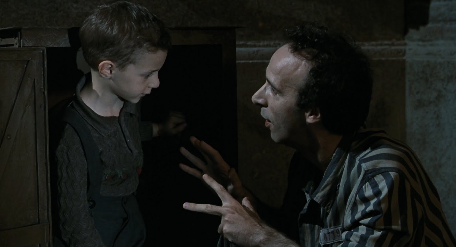 Lindo e lúdico, a grandiosidade do filme está em ter conseguido retratar um tema tão duro e pesado de forma leve, mesmo com momentos de dar nó na garganta.
Guido é um judeu italiano mandando para um campo de concentração, nos anos 40, e lá usa da imaginação para proteger seu filho dos horrores do regime nazista, fazendo-o acreditar que tudo é uma grande competição de brincadeira.
A Vida é Bela levou o prêmio de Melhor Filme Estrangeiro, deixando Central do Brasil a ver navios, e ainda rendeu a Benigni a estatueta de Melhor Roteiro naquele estranho Oscar de 1999.