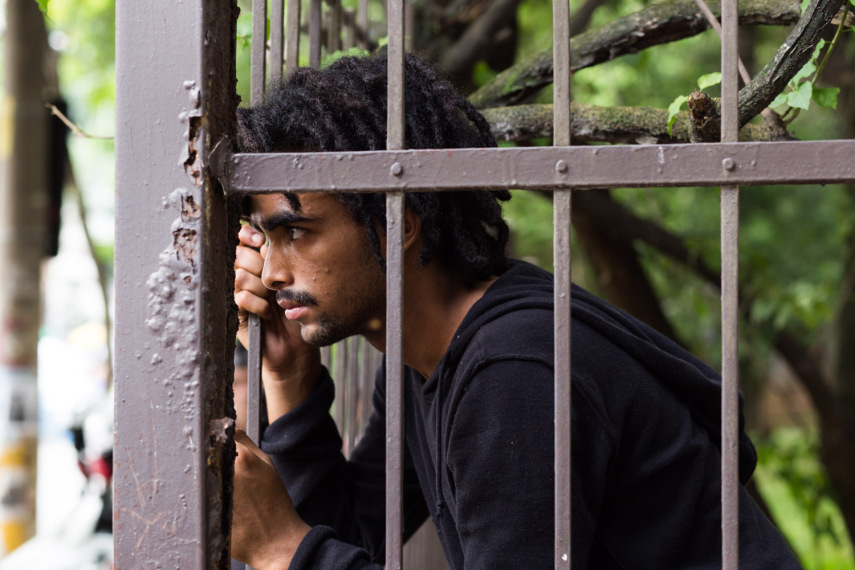 Heudes Cássio da Silva Oliveira é um jovem negro de 18 anos. Ele é morador do Embu, bairro mais afastado do Centro de São Paulo com relação a Pinheiros onde estuda e defende a permanência. Em Embu há muitas atividades artísticas e saraus que entre poesia e canções propõe debates filosóficos e políticos. Apesar da recusa em aceitar o rótulo de líder, já aos 17 anos Heudes era um dos representantes do Movimento Passe Livre (MPL) que incendiou o país com a proposta de gratuidade no transporte. O jovem de voz firme, tranças e chinelos era sempre indicado pelos alunos que chamamos para conversar entre as grades da escola. “Fala com o Heudes primeiro”. É possível que seu pai porteiro e sua mãe que trabalha em um bar não tenham a clareza e a objetividade política de seu tão jovem filho. “Eu acredito que precisamos investir em Educação. Se há bancos e empresas que continuam faturando muito, elas é que têm que pagar a conta da crise”, disse para a gente pendurado nas grades do portão da escola observado por 4 policiais militares com medo que eu chegasse mais perto e entrasse na escola. “A reorganização deve ser conversada com pais, alunos e professores quem sabem os problemas do dia a dia. Aqui mesmo a gente está com privadas quebradas”, conta.