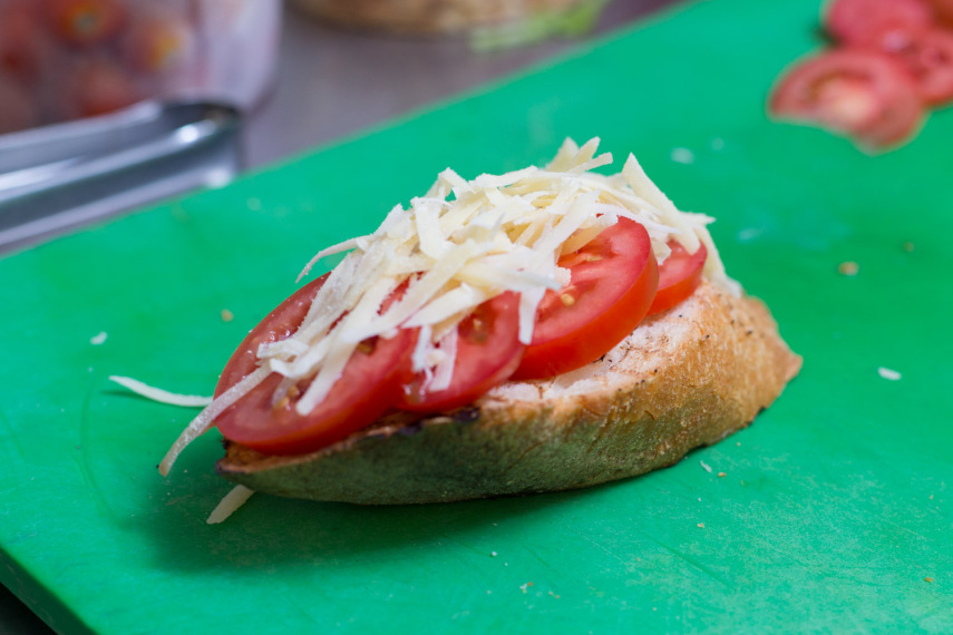 Sem segredos! Pegue uma fatia de pão italiano, passe azeite e doure na chama. Depois, corte fatias de tomate e rale um pouco de parmesão. Monte no pão, exatamente como na foto
